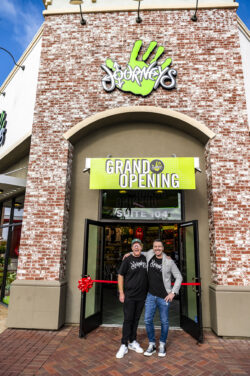 Two men smiling in front of a new journeys store during its grand opening, standing under a sign that reads "grand opening" with a red ribbon, set against a brick facade.