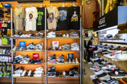 Interior of a clothing and footwear store displaying an array of shoes on lower shelves and colorful graphic tees hanging above. the ambiance is vibrant and well-organized.