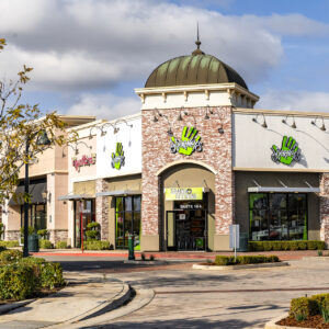 Exterior view of a suburban shopping plaza featuring a store called "hand & stone" with distinctive green palm logo, clear blue sky above, and a neatly paved access road.