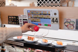 A person places a red ball into a plastic chute beneath an electronic ordering screen at a sushi restaurant. Plates of sushi move along a conveyor belt beneath the screen.