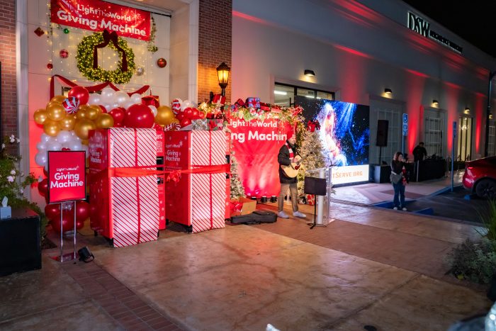A festive outdoor area features large red Giving Machines, holiday decorations, wrapped gift boxes, balloons, a Christmas wreath, and a musician playing guitar near a crowd at night.