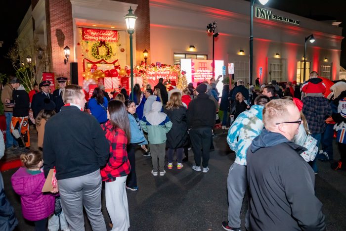 A crowd of people gathers at night outside a brightly lit building decorated with holiday lights and balloons, with a sign reading “Giving Machine” visible in the background.