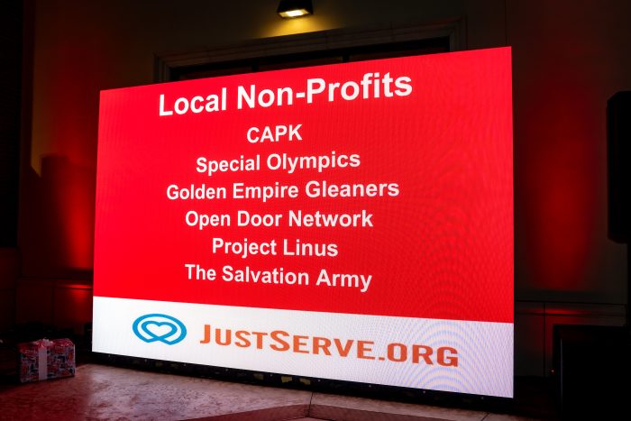 A large digital screen displays a list of local non-profits, including CAPK, Special Olympics, Golden Empire Gleaners, Open Door Network, Project Linus, and The Salvation Army, above the JustServe.org logo.