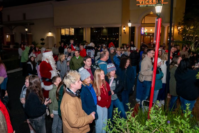A large crowd of people, including children and adults, gathers outdoors at night near a building. Santa Claus is present, interacting with attendees. Some people are dressed warmly, and festive decorations are visible.