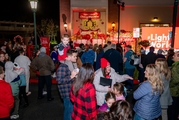 A crowd gathers outside at night near a building decorated with holiday lights and wreaths. A red sign reads "#LightTheWorld." People, including children, are bundled in winter clothes, some smiling and talking.