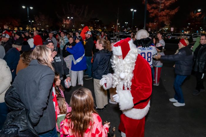A person dressed as Santa Claus interacts with children in a busy outdoor parking lot at night, surrounded by a crowd of people in winter clothing. Holiday lights and trees are visible in the background.