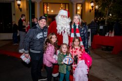 A family with three young girls poses and smiles with a person dressed as Santa Claus outside at night, in front of a lit building decorated for the holidays.