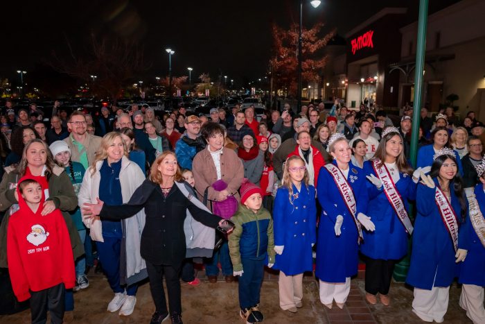 A large crowd gathers outdoors at night, some dressed in festive or winter clothes. Several women in blue coats and sashes stand at the front, smiling, with shops and holiday lights visible in the background.