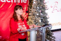 A woman in a red jacket speaks at a clear podium with a microphone. Behind her are holiday lights, a decorated tree, and festive red and white signage. She is smiling and holding papers.