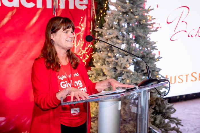 A woman in a red jacket speaks at a clear podium with a microphone. Behind her are holiday lights, a decorated tree, and festive red and white signage. She is smiling and holding papers.