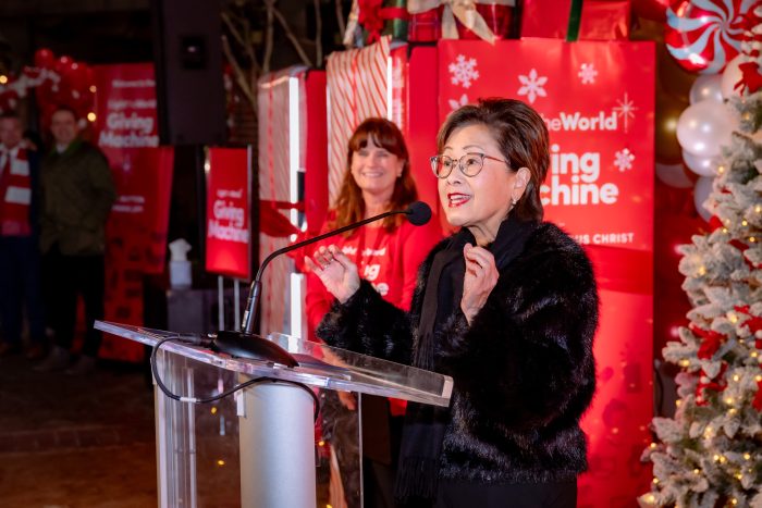 A woman speaks at a podium decorated for Christmas, with a festive backdrop featuring red gift-wrapped boxes and a decorated tree. Another woman stands and smiles in the background.