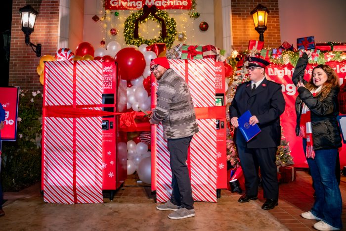 A man in a red hat cuts a large red ribbon on two festive red-and-white vending machines, surrounded by balloons and holiday decorations, while two people stand nearby smiling.