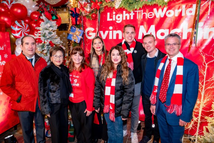 A group of eight people, some wearing red and white scarves, pose and smile in front of a festive holiday display with Christmas decorations and a sign that reads “Light The World Giving Machine.”.