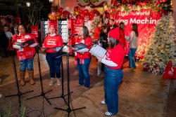 Four women in red "Giving Machine" shirts sing at microphones in a festively decorated area with Christmas trees, lights, and Giving Machines. People in the background observe the performance. The atmosphere is cheerful and holiday-themed.