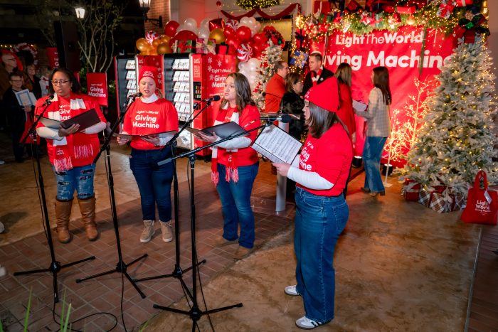 Four women in red "Giving Machine" shirts sing at microphones in a festively decorated area with Christmas trees, lights, and Giving Machines. People in the background observe the performance. The atmosphere is cheerful and holiday-themed.
