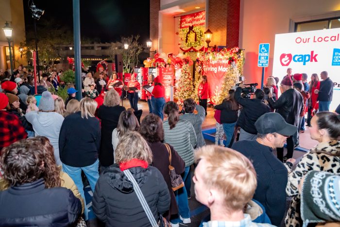 A crowd gathers outdoors at night for a holiday event with festive decorations, red banners, and a stage where people in red shirts are speaking. Some attendees are filming or taking photos.