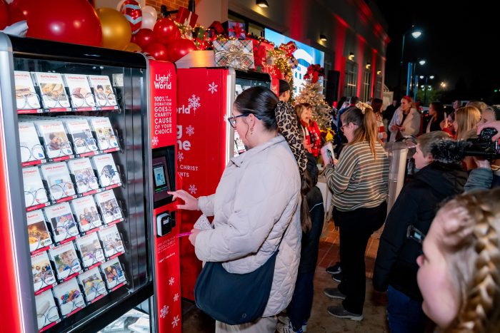 People gather outdoors at a brightly lit event, using red vending machines labeled "Light the World." The machines are filled with donation cards, and festive holiday decor like balloons and garlands is visible above.