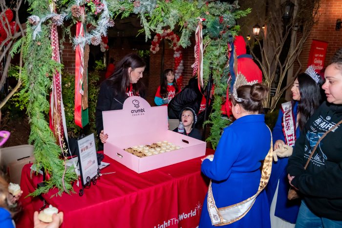 A woman serves cookies from a pink Crumbl Cookies box at a festive outdoor booth decorated with greenery and ornaments. Several people, including children and a woman in a crown and sash, wait in line.