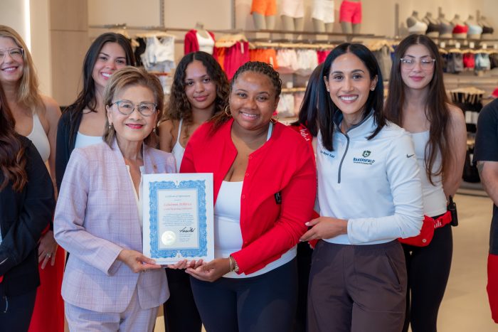 A group of women stand together smiling; the woman in front on the left holds a framed certificate while two others stand close, also smiling, with athletic wear displayed in the background.