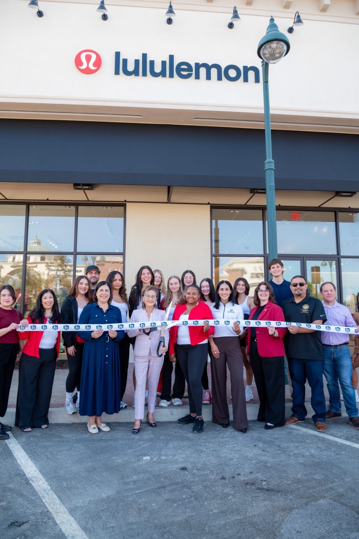 A group of people stands outside a Lululemon store for a ribbon-cutting ceremony, smiling for the photo. The storefront and Lululemon logo are visible above them.