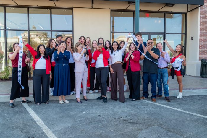 A group of people, some holding scissors, gathers outside a building for a ribbon-cutting ceremony. Most are smiling and cheering, with a mix of business and casual attire.