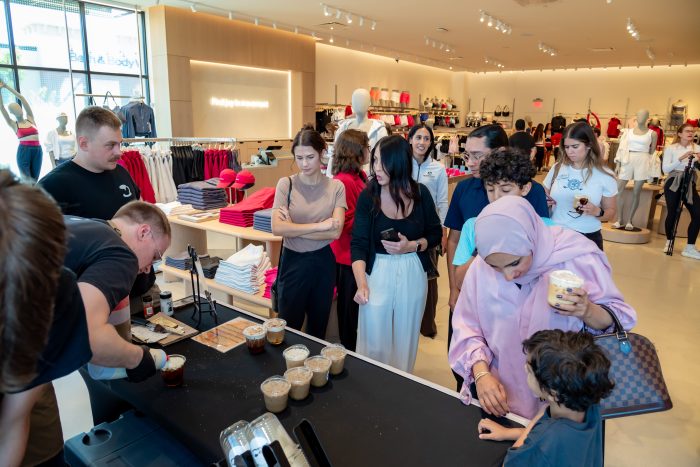 People gather around a table with drinks and samples in a bright clothing store. Various shoppers, including adults and kids, interact and watch as two workers prepare beverages. Clothing displays are visible in the background.