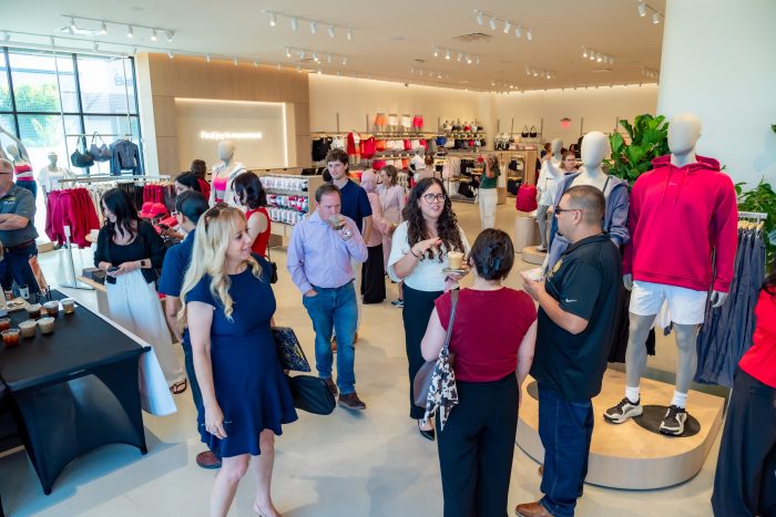 People mingle and chat at a clothing store event. Mannequins display sporty outfits, and racks of clothes line the walls. Bright lighting and a modern interior create a welcoming atmosphere.