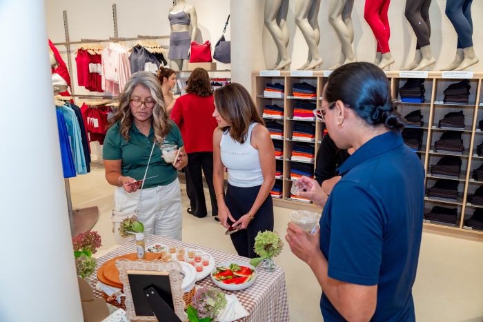 Four people gather around a small table with snacks and drinks inside a retail store selling athletic clothing, with shelves of folded clothes and mannequins in the background.