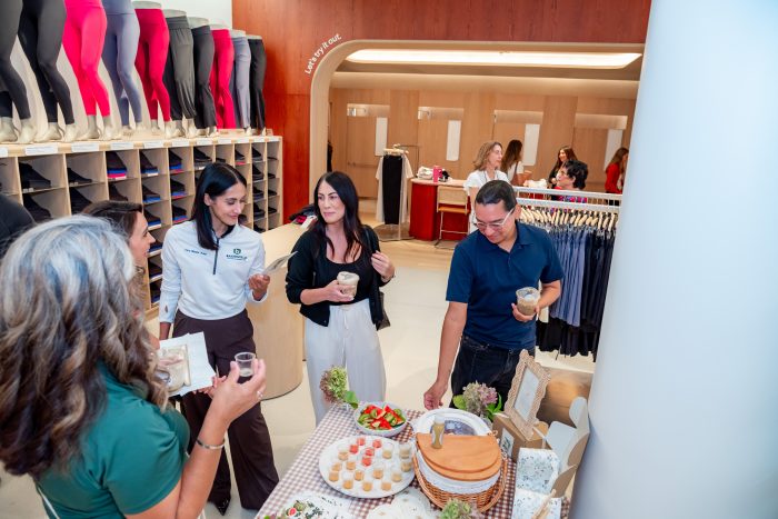 Four people stand around a table with food samples at a clothing store, talking and holding drinks. Mannequin legs in leggings line the shelves in the background.