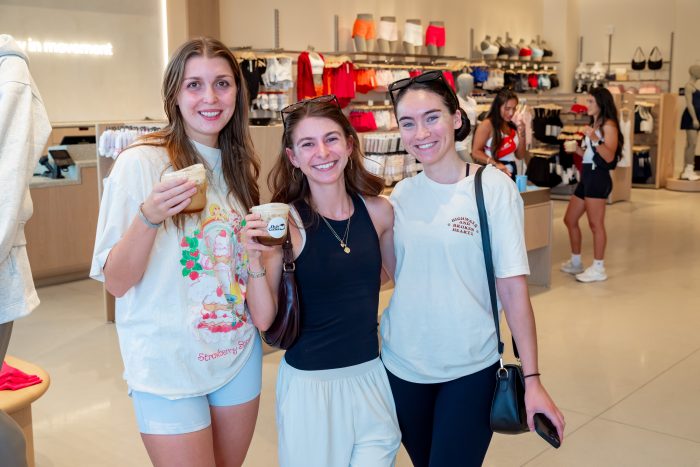 Three women stand together smiling in a clothing store, each holding a drink. Athletic wear is displayed on racks and shelves behind them. Other shoppers are visible in the background.
