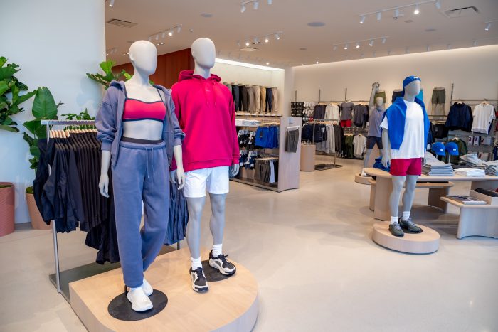 Three mannequins in sporty outfits are displayed in a modern, well-lit clothing store. Racks of casual and athletic wear line the walls, and green plants add a touch of decor.