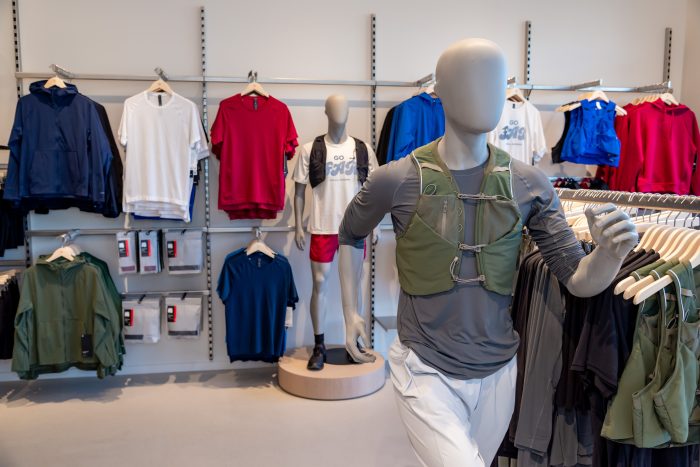 A running mannequin dressed in athletic gear is in the foreground. Behind it are racks and shelves displaying t-shirts, shorts, and jackets in various colors in a sports clothing store.