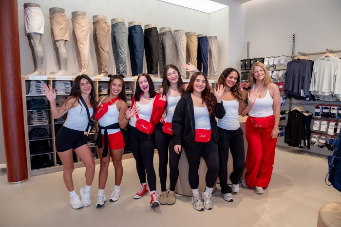 Seven women in athletic wear stand smiling and waving inside a clothing store, with shelves of neatly folded pants and shorts displayed in the background.