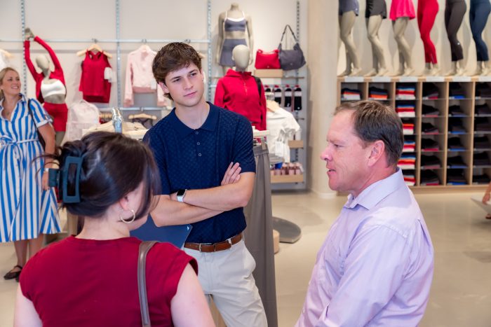 Three people have a conversation inside a clothing store, with folded shirts and mannequins in the background. One woman in a striped dress stands off to the side, smiling.