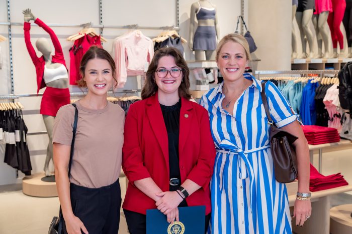 Three women smiling and standing together in a clothing store, with activewear displayed in the background. One woman is holding a blue folder with a gold emblem.