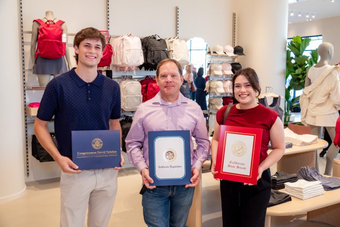 Three people stand indoors, each holding a certificate or folder with official seals. Behind them are shelves displaying bags, clothes, and accessories in a brightly lit retail store.