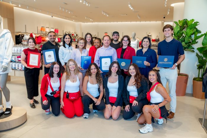 A group of people pose together in a modern, bright store, with some holding certificates and folders. Several women in red and white athletic outfits kneel and stand in front, smiling. Mannequins and plants are visible in the background.