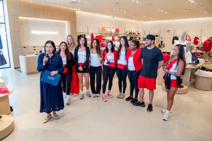 A woman speaks holding a blue folder while standing beside a group of people dressed in coordinated red, white, and black outfits inside a bright, modern clothing store.